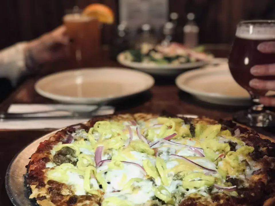 A pizza topped with cheese, pickles, onions, and possibly other ingredients, on a wooden table in a restaurant setting.