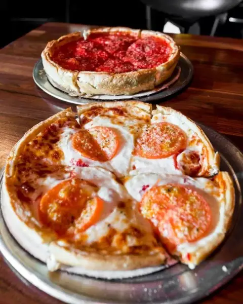 Two pizzas on metal trays, one with pepperoni and the other with cheese and pepper slices, on a wooden table.